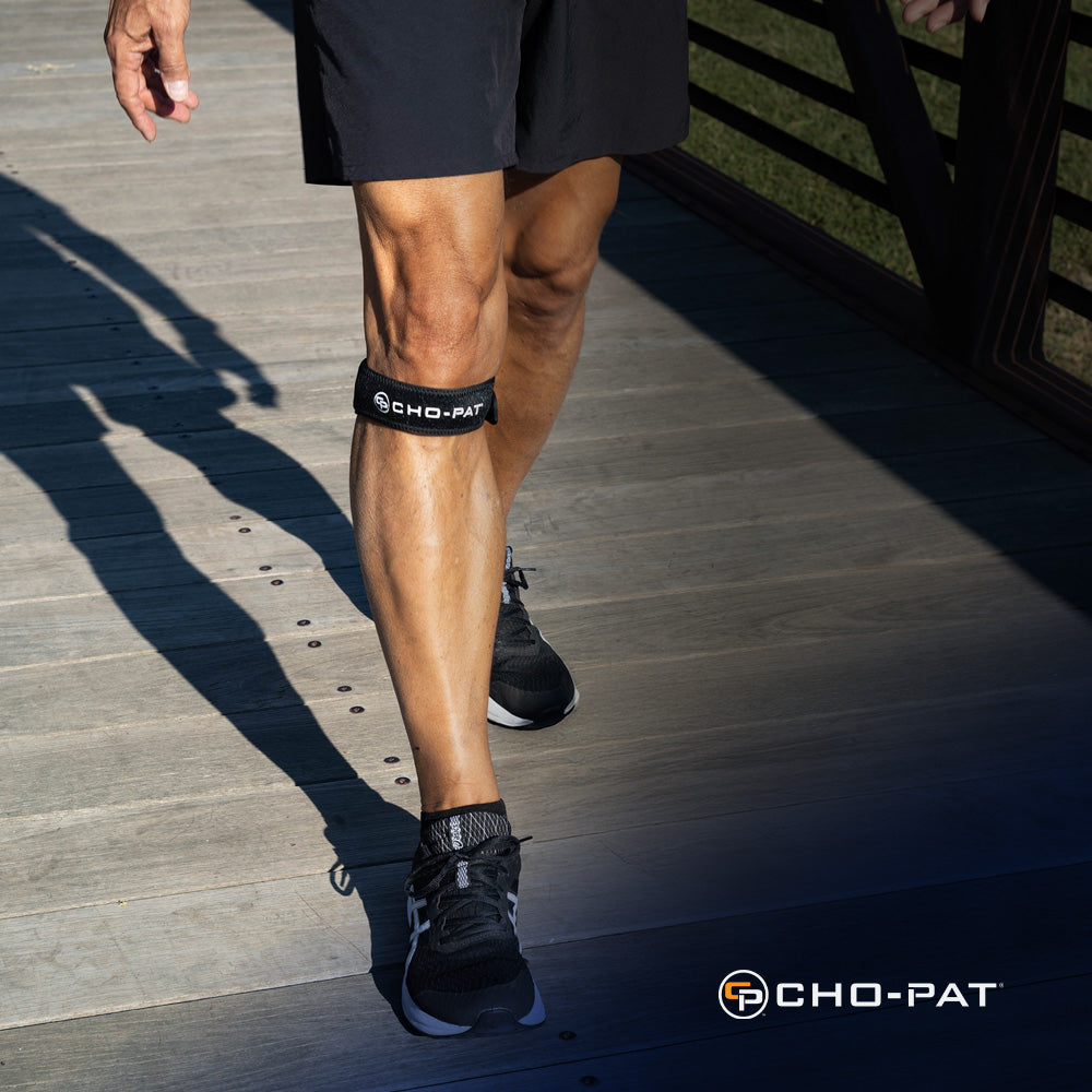 Person walking outdoors on a wooden bridge while wearing the Cho-Pat Patella Knee Strap below the kneecap.